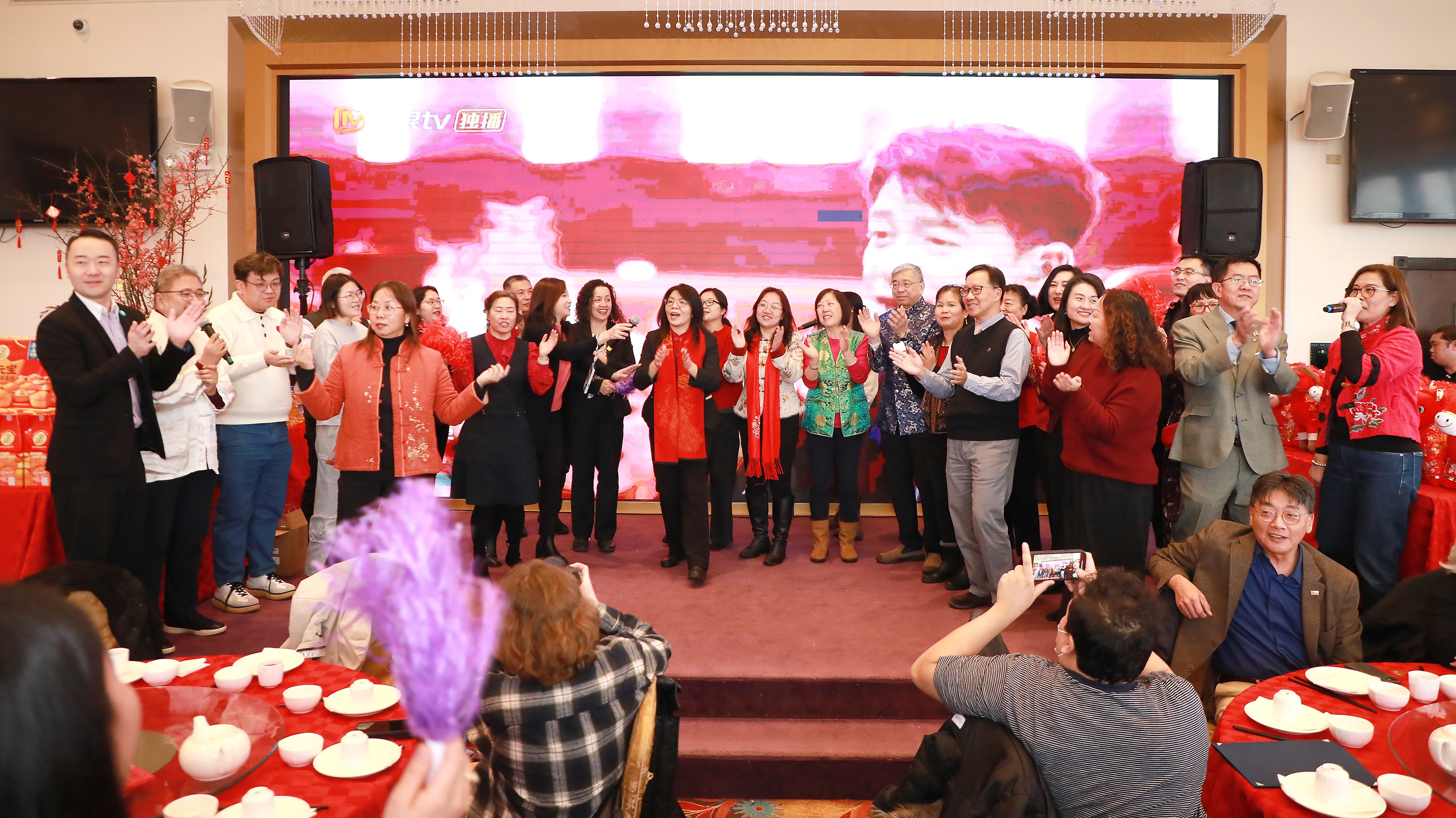 一群人在庆祝活动中唱歌和欢呼，周围有节日装饰，背景是一个大屏幕。